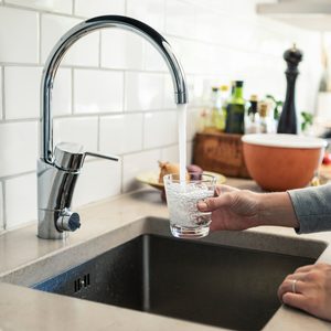 A hand holds a glass under a shiny faucet, filling it with water in a kitchen with a bowl and bottles in the background.