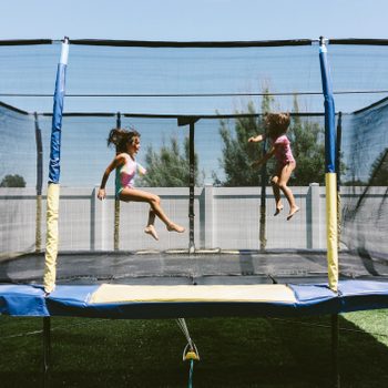 Two children jump on a trampoline in a sunny backyard, surrounded by a fence and green grass, enjoying playtime in summer attire.
