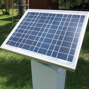 A solar panel captures sunlight, mounted on a pole amidst green grass and trees, providing a sustainable energy source in a natural setting.