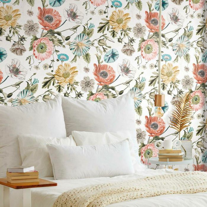 A bed with white pillows rests against a floral-patterned wall, accompanied by a side table with books, and a decorative plant beneath a pendant light.