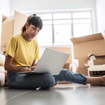 A woman sits on the floor, using a laptop amidst unpacked boxes. Natural light from a window illuminates the cluttered, yet bright room.