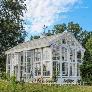 A white greenhouse stands with open doors, showcasing plants inside, surrounded by tall grass and trees under a blue sky with scattered clouds.