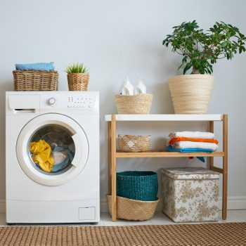 A washing machine displays colorful laundry. A shelf beside holds neatly folded towels and decorative baskets, set against a light wall with a potted plant.
