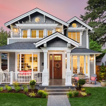 A two-story, gray-shingled house with white trim stands in a landscaped yard, featuring a welcoming porch and large windows, surrounded by trees at sunset.