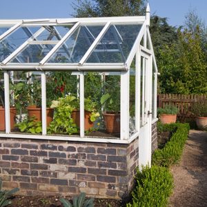 A glass greenhouse filled with potted plants stands on a brick base, surrounded by a manicured garden with a wooden fence in the background.