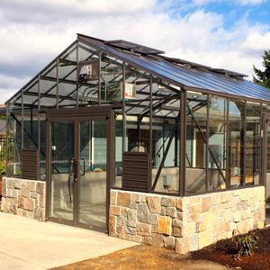 A glass greenhouse stands with stone accents, featuring sliding doors and a sloped roof, surrounded by grass and a clear blue sky.