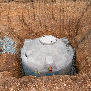 A concrete tank is partially buried in a dug-out area, surrounded by gravel and soil, indicating preparation for installation.