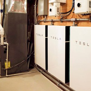 Three Tesla battery energy storage units are mounted on a wall, connected to equipment in a basement-like space, alongside pipes and a HVAC unit.