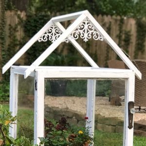 A small, white greenhouse with a decorative roof contains various plants, situated on lush grass near a stone path and outdoor seating area.
