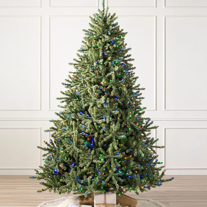 A decorated Christmas tree stands upright, illuminated with colorful lights, placed on a white rug in front of a light-colored wall.