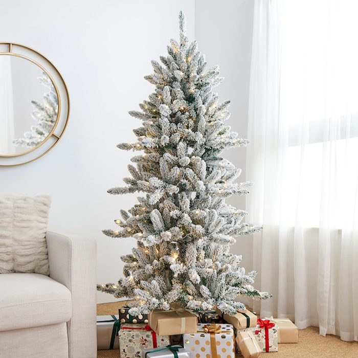 A pre-lit, frosted Christmas tree stands beside a couch, surrounded by wrapped gifts, with a mirror reflecting its image, in a softly lit room.