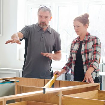 A man gestures while discussing cabinetry with a woman, who measures with a tape in a bright, unfinished kitchen space with windows.
