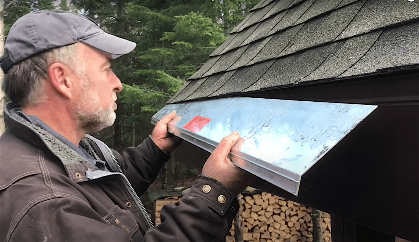 A man holds a metal flashing against a roof edge in a wooded area, while a stack of logs is visible in the background.