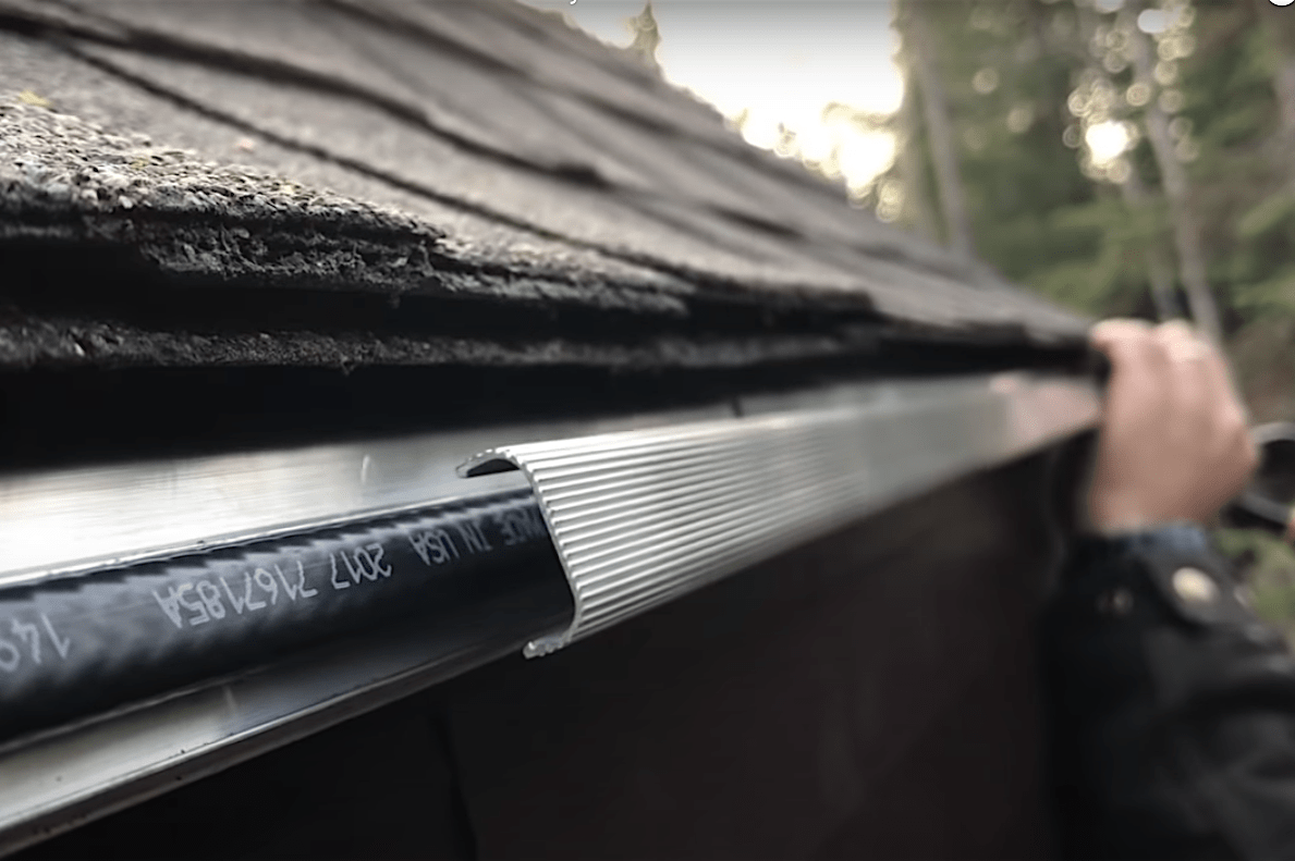 A person is installing a guard on a roof's edge, enhancing water flow management in a wooded environment with a blurred background of trees.