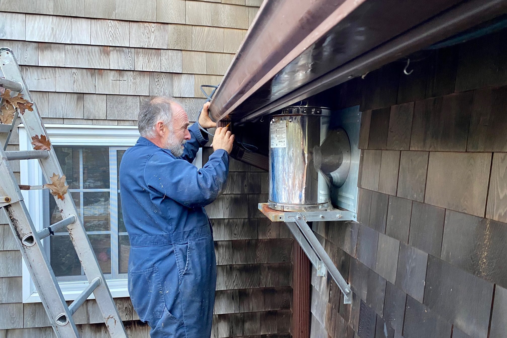 A man adjusts a metal vent while standing on a ladder next to a wooden shingle wall, working on home maintenance outdoors.