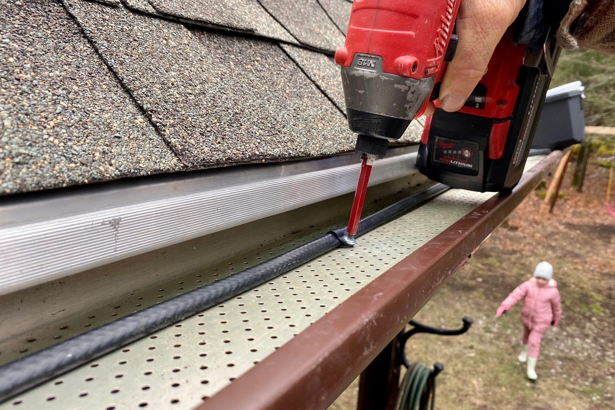 A hand uses a power drill to attach a fitting to a black hose along a metal gutter, while a child walks in the background.
