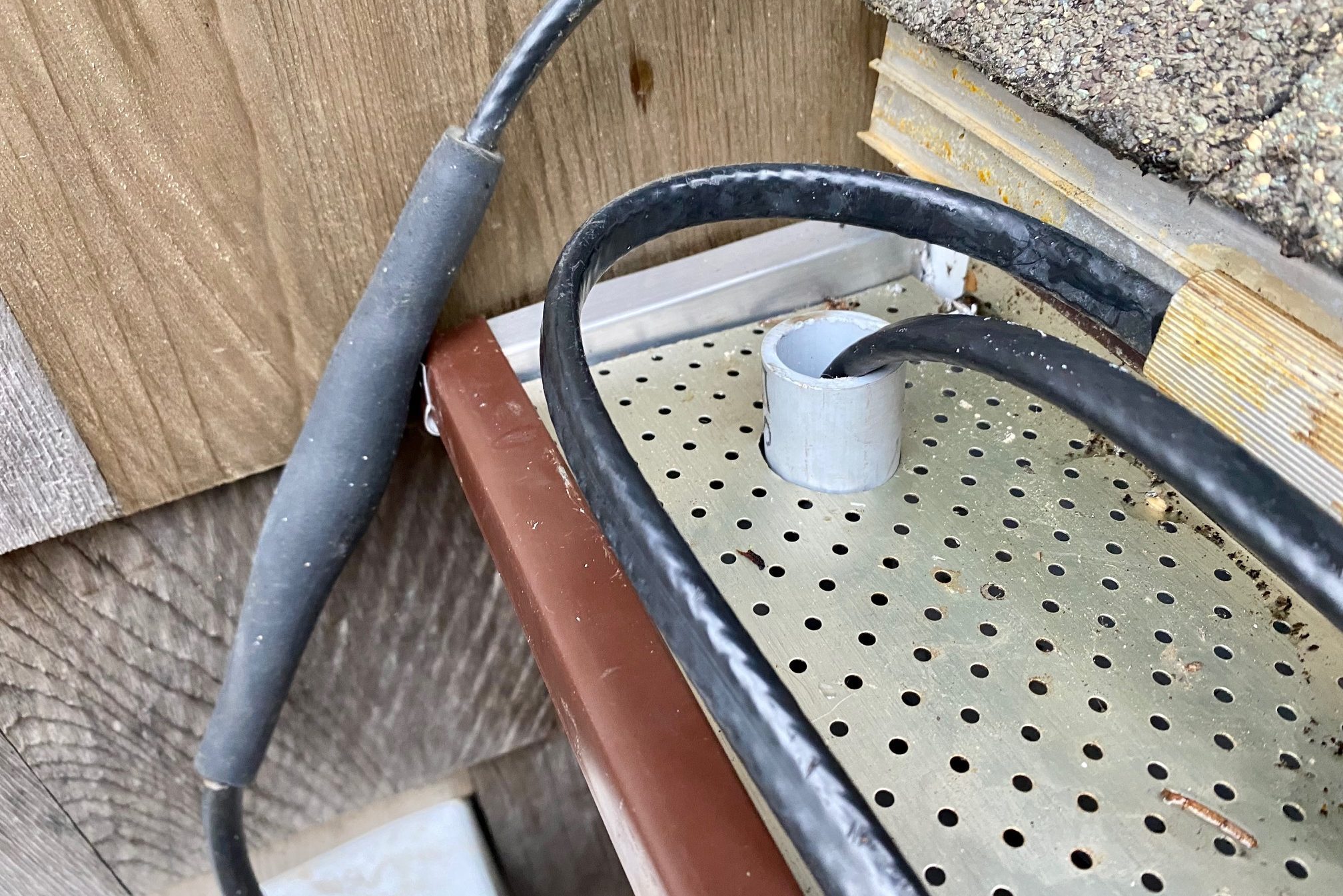 A black cable lies on a perforated metal surface, connecting to a gray pipe fitting, situated against a wooden wall. Debris surrounds the area.