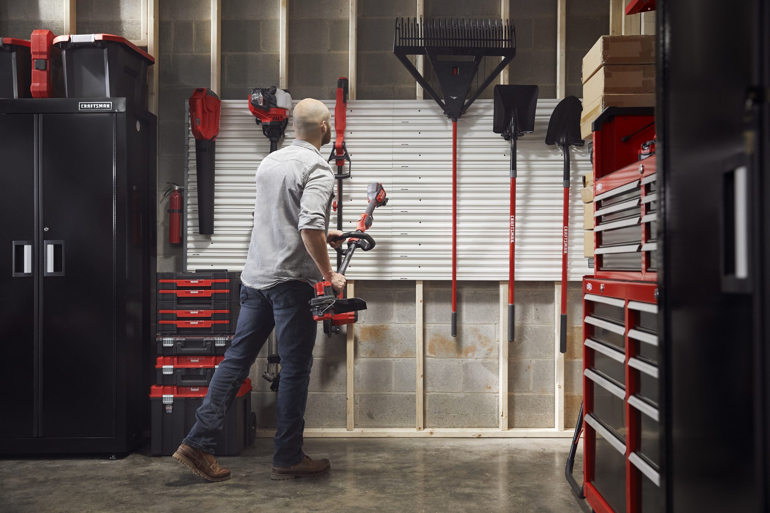 A person holds a power tool while standing in a garage filled with organized tools, cabinets, and equipment along a concrete wall.