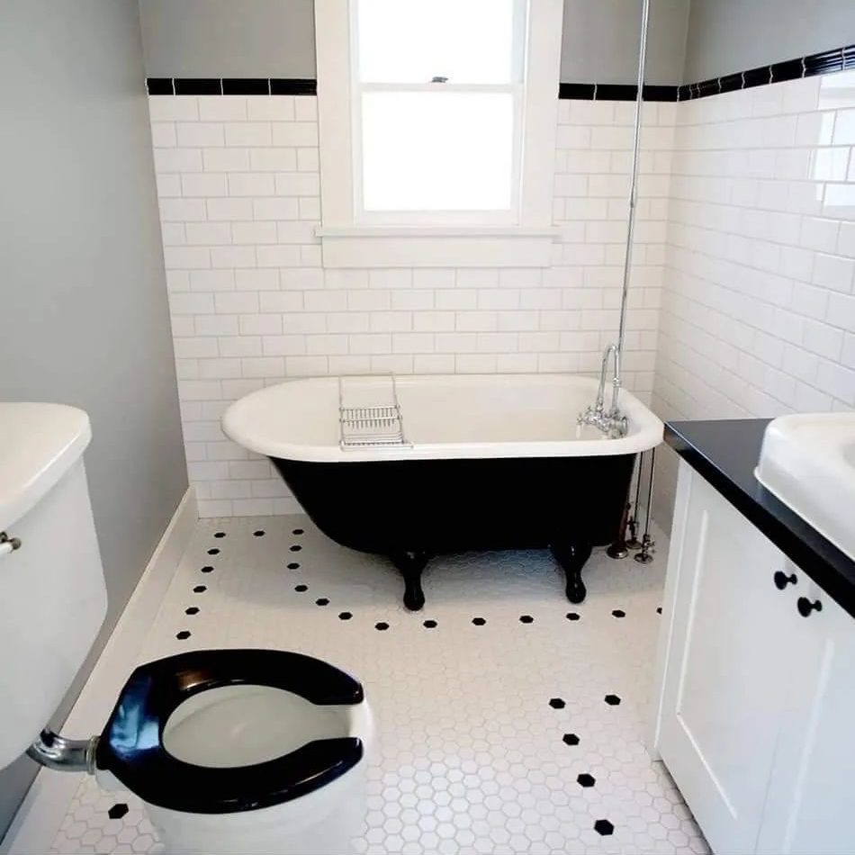 A black and white clawfoot bathtub sits beside a toilet in a modern bathroom with hexagonal tiles and white subway tile on the walls.