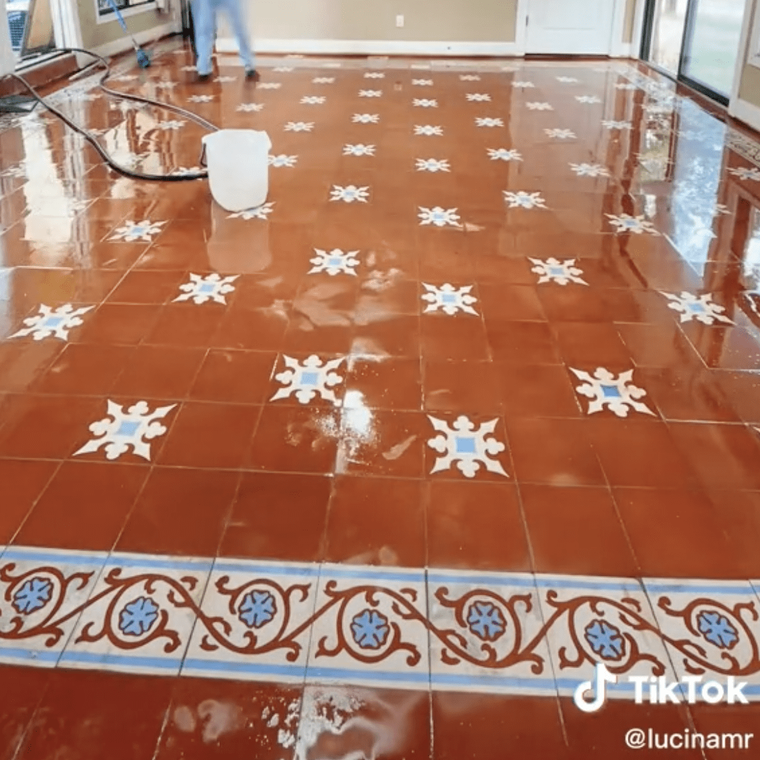 A person is cleaning a shiny, tiled floor featuring intricate designs, in a well-lit room with natural light coming through windows.