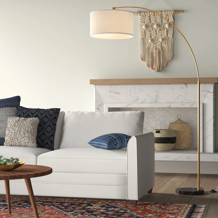 A white sectional sofa with pillows sits beside a curved floor lamp, on a colorful rug, near a marble fireplace adorned with woven decor.