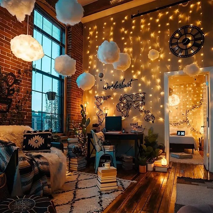 A cozy room features books stacked beside a desk under whimsical, cloud-like decorations and string lights illuminating a map and 