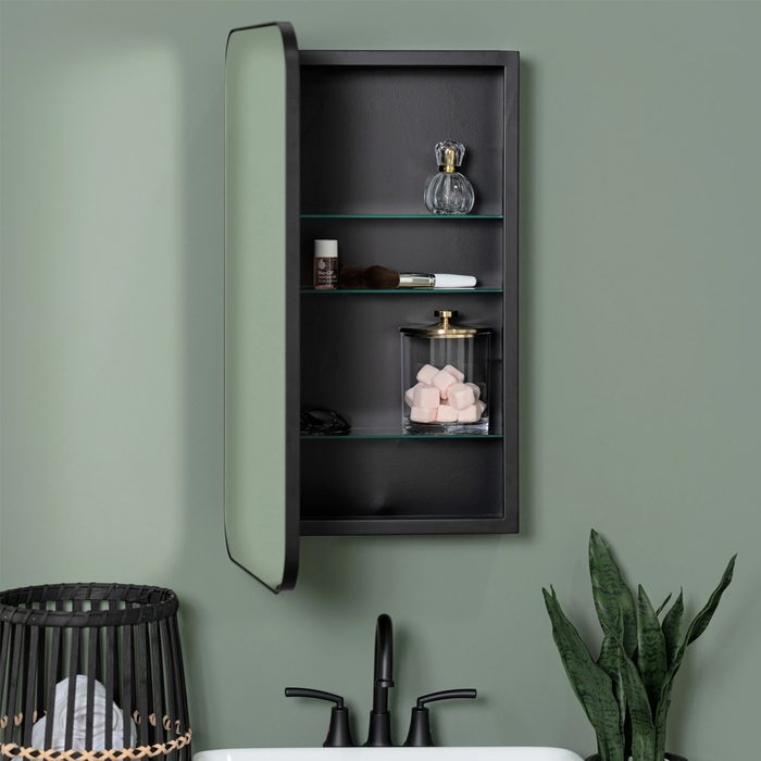 A wall-mounted cabinet holds cosmetics and toiletries, while a black faucet and a dark basket accompany a green wall and indoor plant in the bathroom.