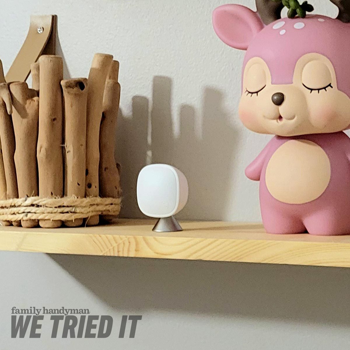 A white device stands on a wooden shelf beside a pink cartoon deer figurine and a basket made of wooden sticks, against a light wall.