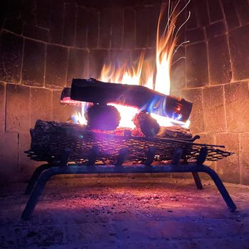 A fire burns brightly on a metal grate, surrounded by stone walls, casting warm light and flickering shadows in a cozy setting.