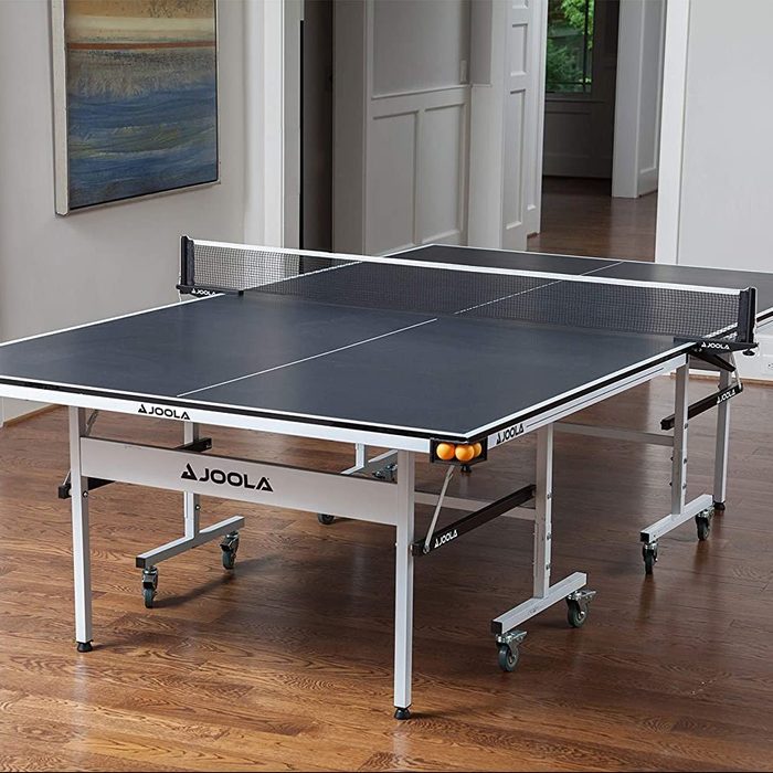 A ping pong table stands in a well-lit room with wooden flooring, featuring a net in the center and orange balls near one corner.