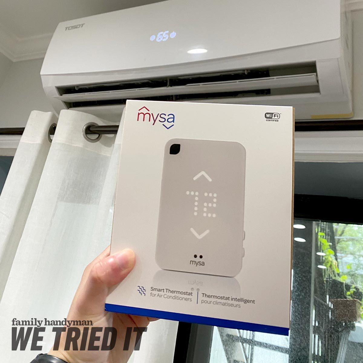 A person holds a smart thermostat box labeled "mysa" in front of an air conditioning unit displaying the temperature, with curtains and a window nearby.