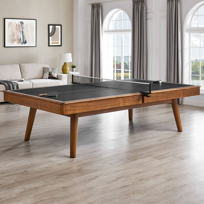 A wooden ping pong table with a black playing surface and net stands in a well-lit, modern living room featuring windows, a sofa, and decor.