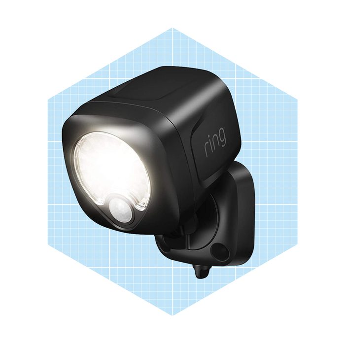 A black outdoor security light is mounted on a wall, illuminating brightly while integrating a motion sensor, set against a light blue grid background.