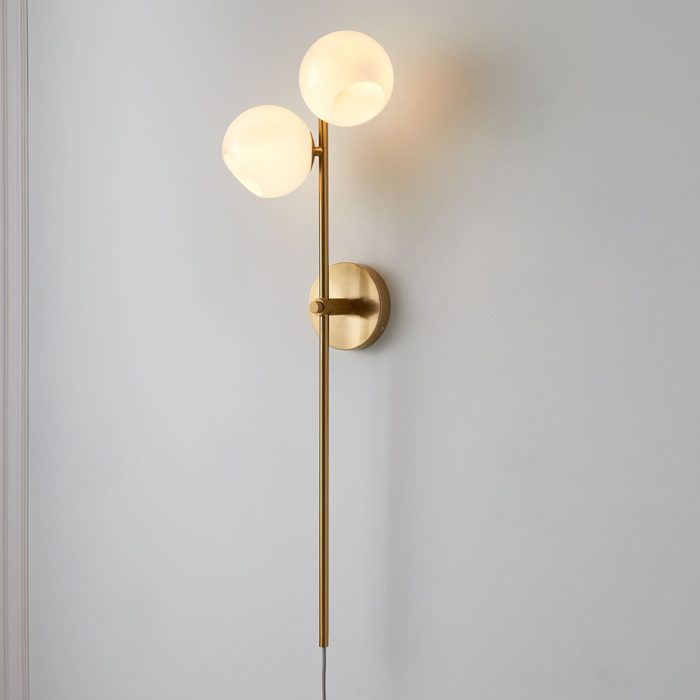 A sleek wall lamp with two round, frosted glass globes emits warm light, mounted on a simple brass fixture against a plain wall.