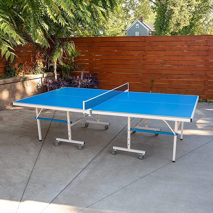 A blue ping pong table is set up outdoors, featuring a net dividing the two halves, surrounded by greenery and a wooden fence.