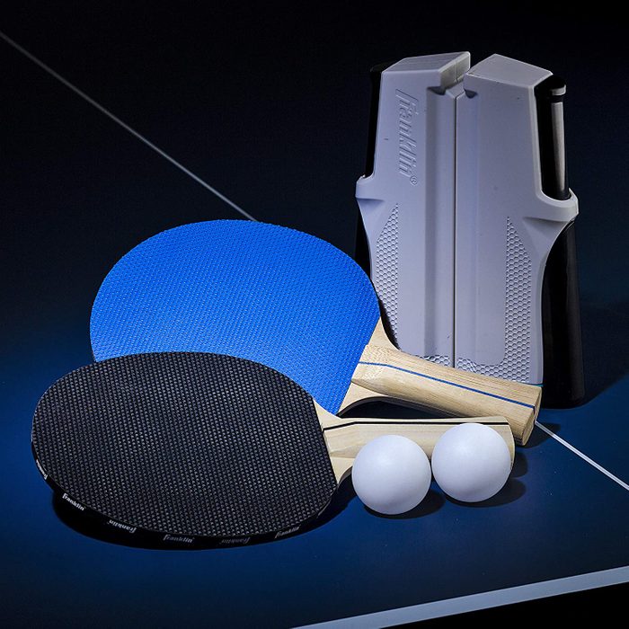 Two table tennis paddles with blue and black surfaces rest next to two white balls, alongside a paddle holder on a dark table.