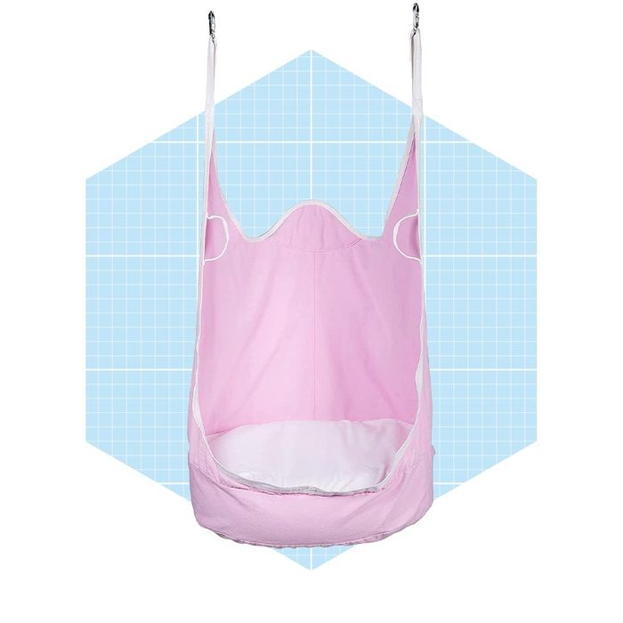 A pink fabric baby hammock hangs suspended, featuring white trim. It's set against a light blue, grid-patterned background, suggesting a playful, soft atmosphere.