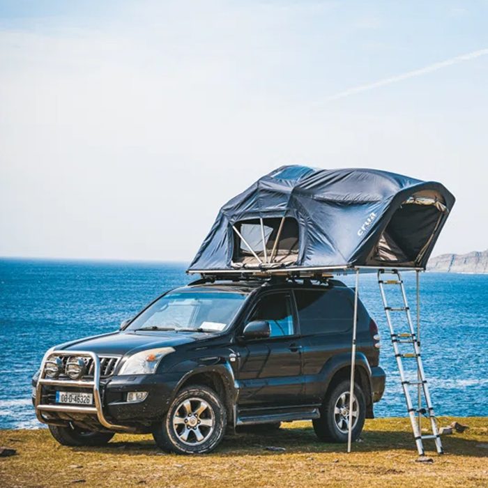 A black SUV with a rooftop tent is parked near the ocean, supported by a ladder, with a clear sky and coastline in the background.