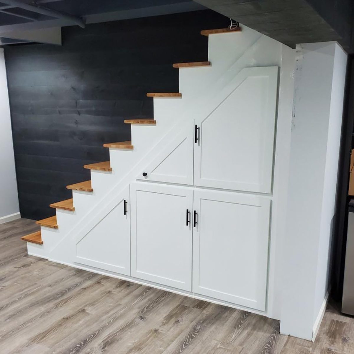 A staircase with wooden steps ascends against a dark wall; beneath, there are white cabinets with black handles, set on a wooden floor.