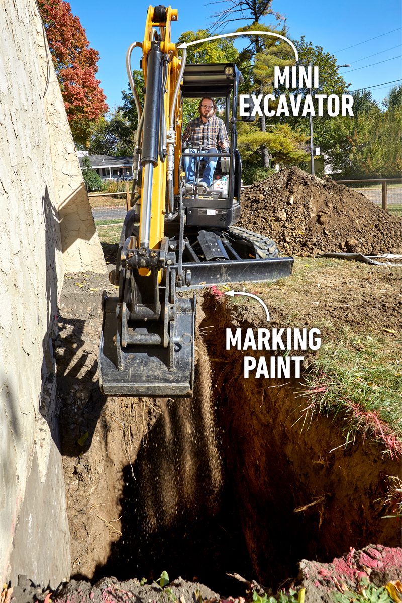 A mini excavator digs into the ground beside a building, with dirt being displaced. Nearby, there is marked paint outlining an area for excavation.