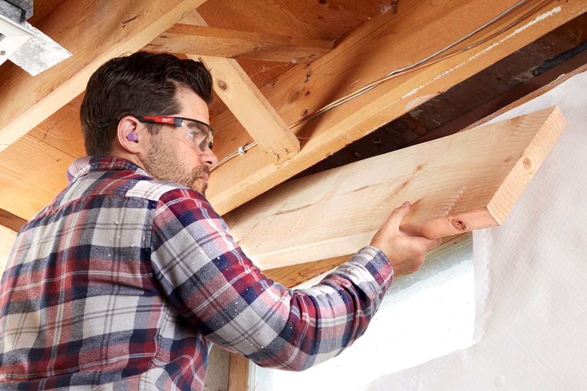 A man wearing safety glasses lifts a wooden beam into place within a wooden frame, working in a construction setting with visible insulation and beams.