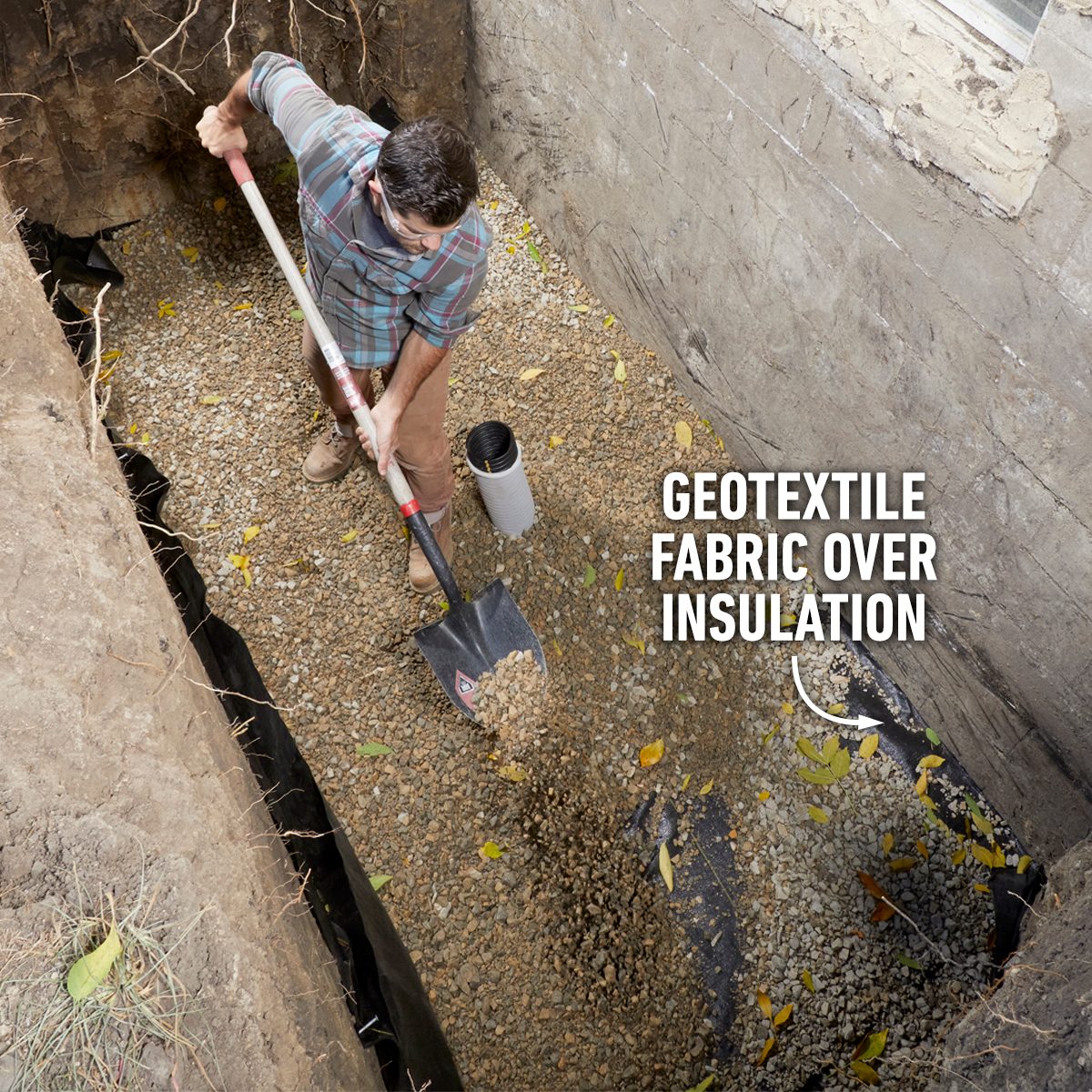 A person shovels gravel into a pit, covering insulation with geotextile fabric, surrounded by rough earth and a dark wall of the excavation.