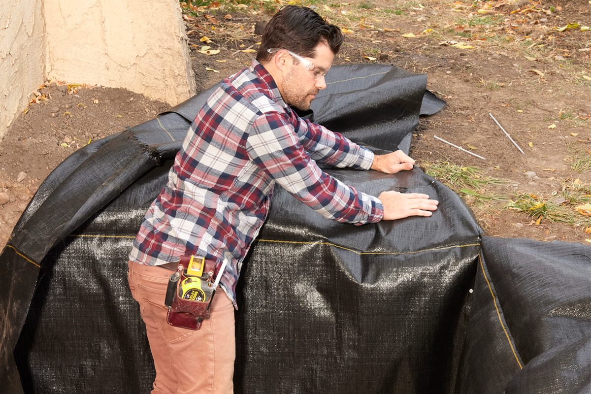 A man in plaid leans over black landscaping fabric, preparing it in an outdoor space surrounded by dirt and scattered leaves.