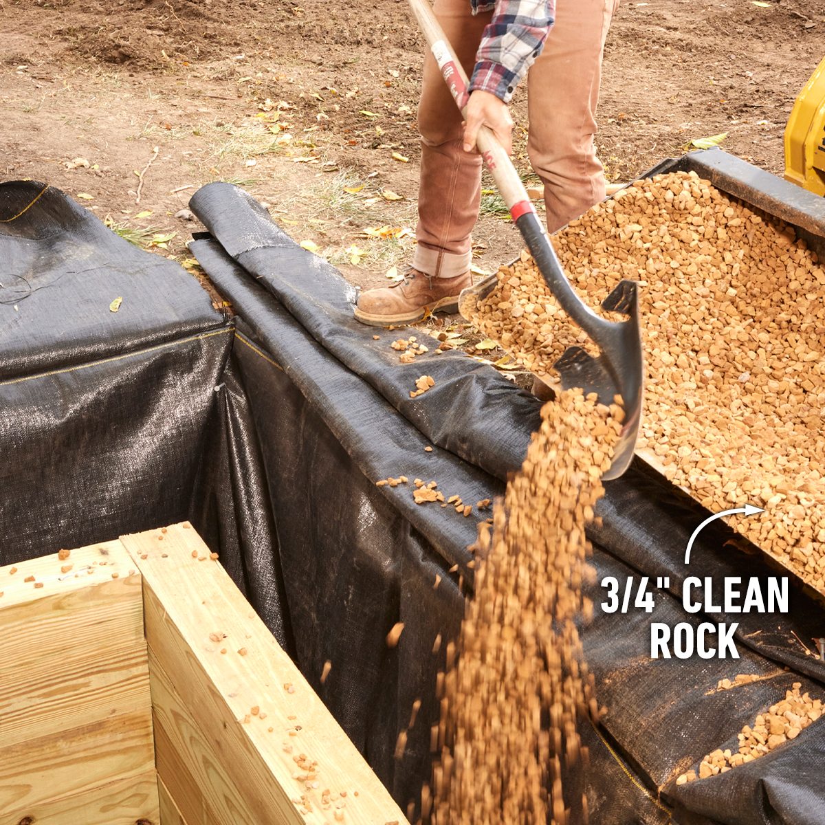 A person shovels clean rock into a black fabric-lined area, with wooden edges visible, in an outdoor construction site.