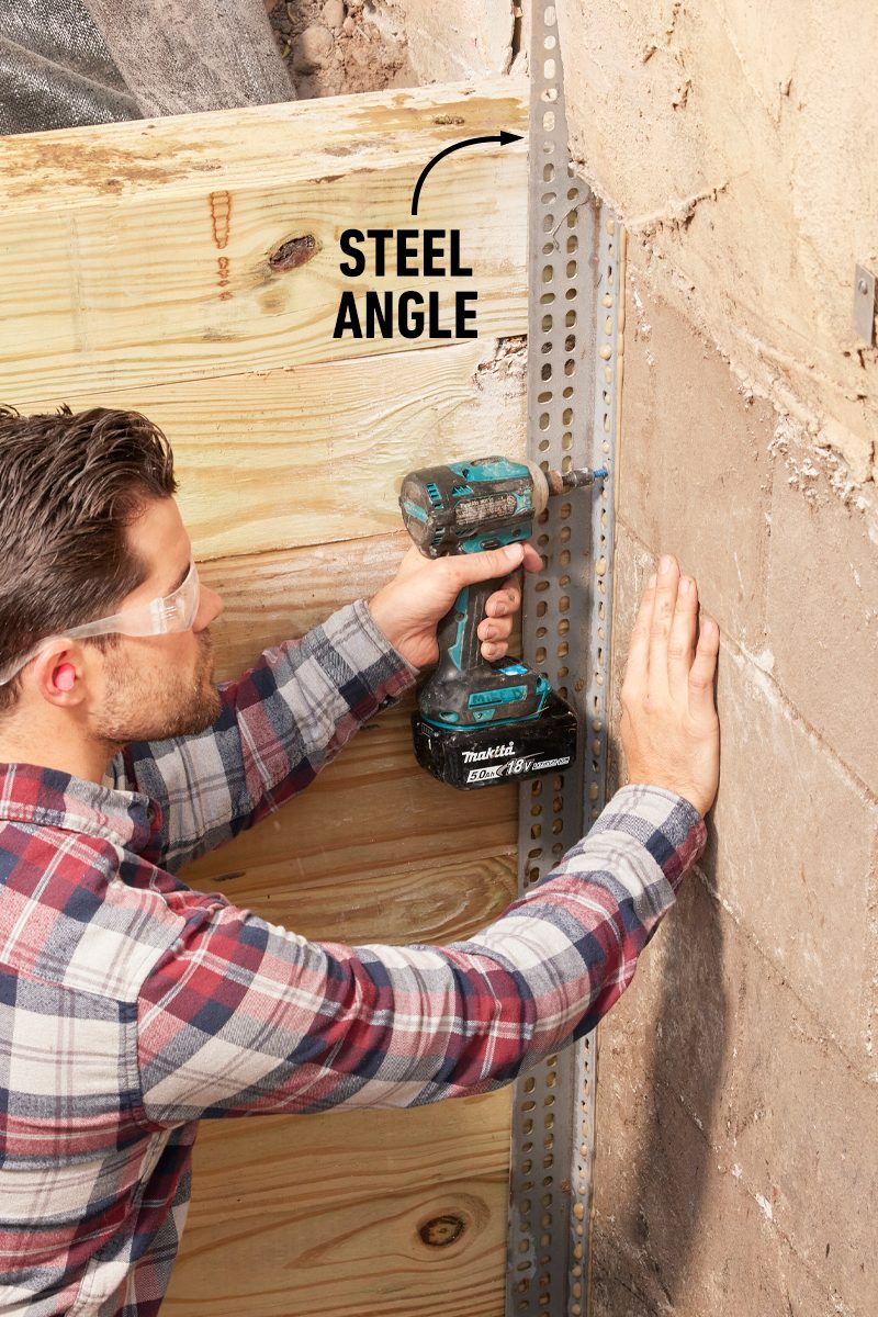 A man uses a cordless drill to attach a steel angle to a wall next to wooden planks, wearing safety glasses and ear protection.