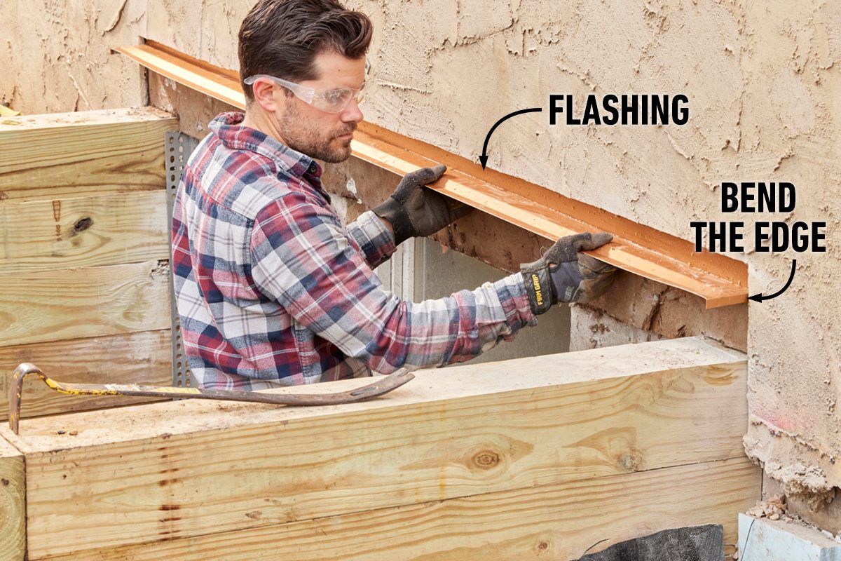 A man in safety glasses installs copper flashing on a wall, using gloves to hold and bend it, surrounded by wooden structures and a textured surface.