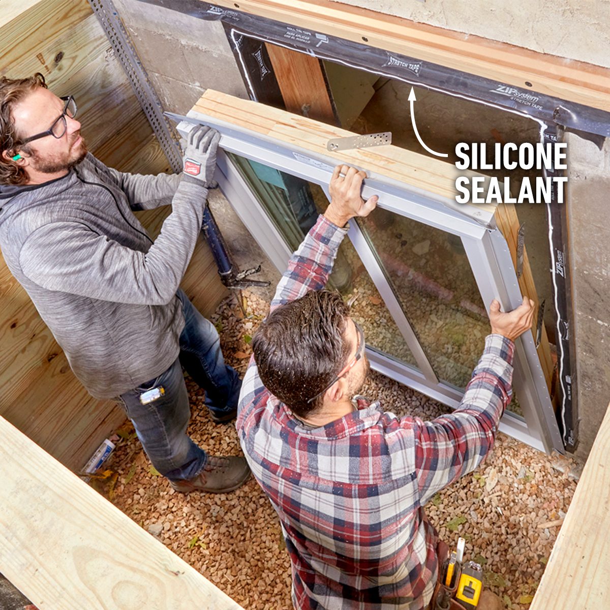 Two men install a window frame, applying silicone sealant. They work in a gravel-filled, semi-enclosed area, surrounded by wooden structures.