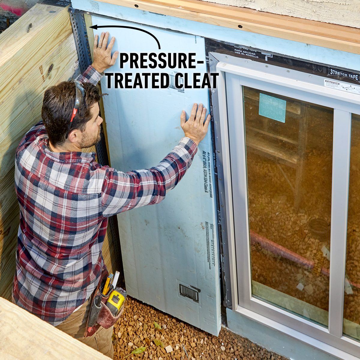 A worker installs a pressure-treated cleat against insulation on a wall near a window, surrounded by wooden structures and gravel flooring.