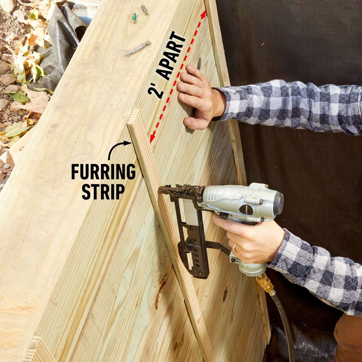 A hand holds a nail gun, attaching furring strips spaced two feet apart on a wooden surface, surrounded by debris and a dark backdrop.