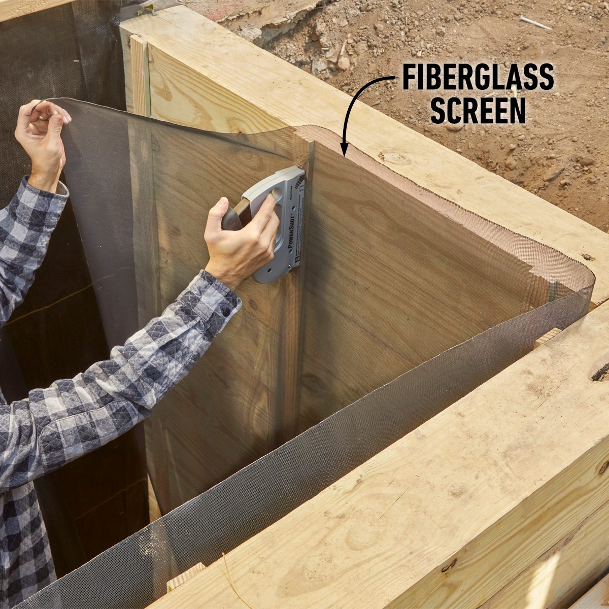 A person is attaching a fiberglass screen to wooden framing using a stapler, positioned above an excavated area with dirt and wooden surfaces surrounding them.